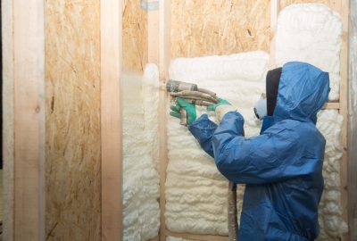 A worker in protective blue coveralls and a respirator applying spray foam insulation to wooden wall studs.