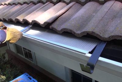 Gloved hands installing a metal mesh gutter guard under a clay tile roof.