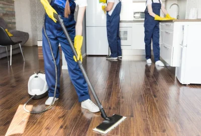 Professional cleaners in blue overalls and yellow gloves cleaning a modern home's wood floors and kitchen.