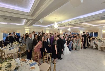Group of wedding guests in formal wear posing in a brightly lit ballroom with marble floors.