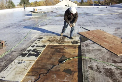 Worker in a hard hat using a power drill on a flat roof with exposed plywood sections.