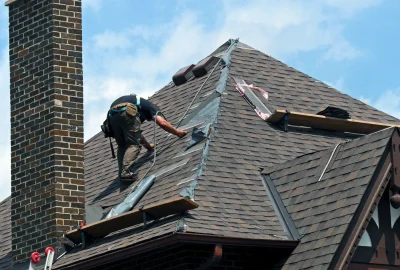 A roofer in a safety harness works on a steep shingled roof next to a brick chimney.