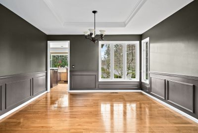 Empty dining room with dark gray wainscoted walls, polished hardwood floors, a chandelier, and large windows.