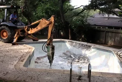 An excavator with a jackhammer attachment breaks up a concrete swimming pool in a backyard.