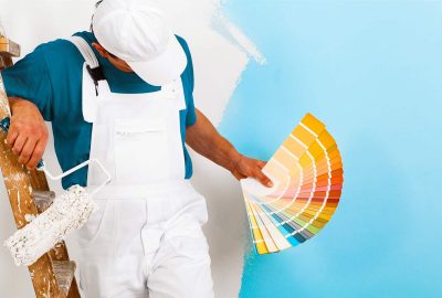 Painter in white coveralls holds a paint roller and a color swatch fan against a partially painted blue wall.
