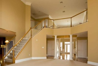 Spacious two-story foyer with tan walls, carpeted stairs, a white railing, and decorative columns near the front entrance.