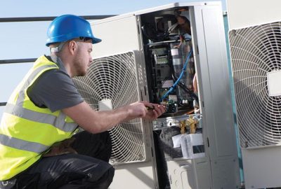 Technician in a safety vest and hard hat repairing an outdoor HVAC unit.