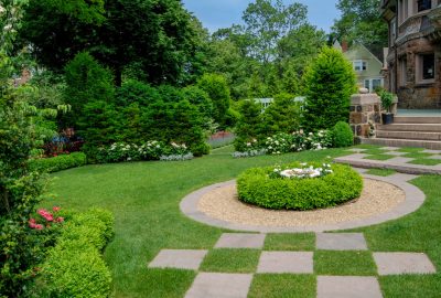 Lush green backyard with a circular gravel garden bed, stone checkered walkway, and mature trees near a stone house.