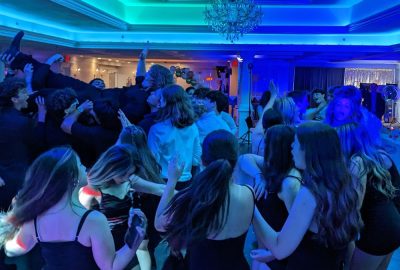 A crowd of teenagers dancing at an event under blue lighting, with a group hoisting a person up in the air.