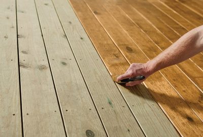 Hand using a brush to stain unfinished wooden deck planks a warm brown color.