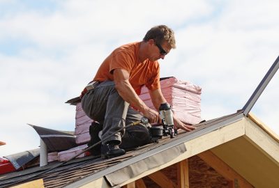 A construction worker in an orange shirt uses a pneumatic nail gun to install shingles on a roof.