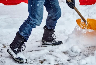 Person in blue jeans and black winter boots shoveling snow with a yellow shovel.