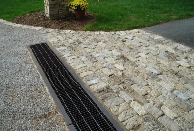 Long black metal drainage grate separating a gravel area from a cobblestone walkway.