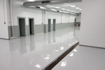 Large, empty industrial room with shiny gray epoxy flooring, white walls, and visible overhead ductwork.