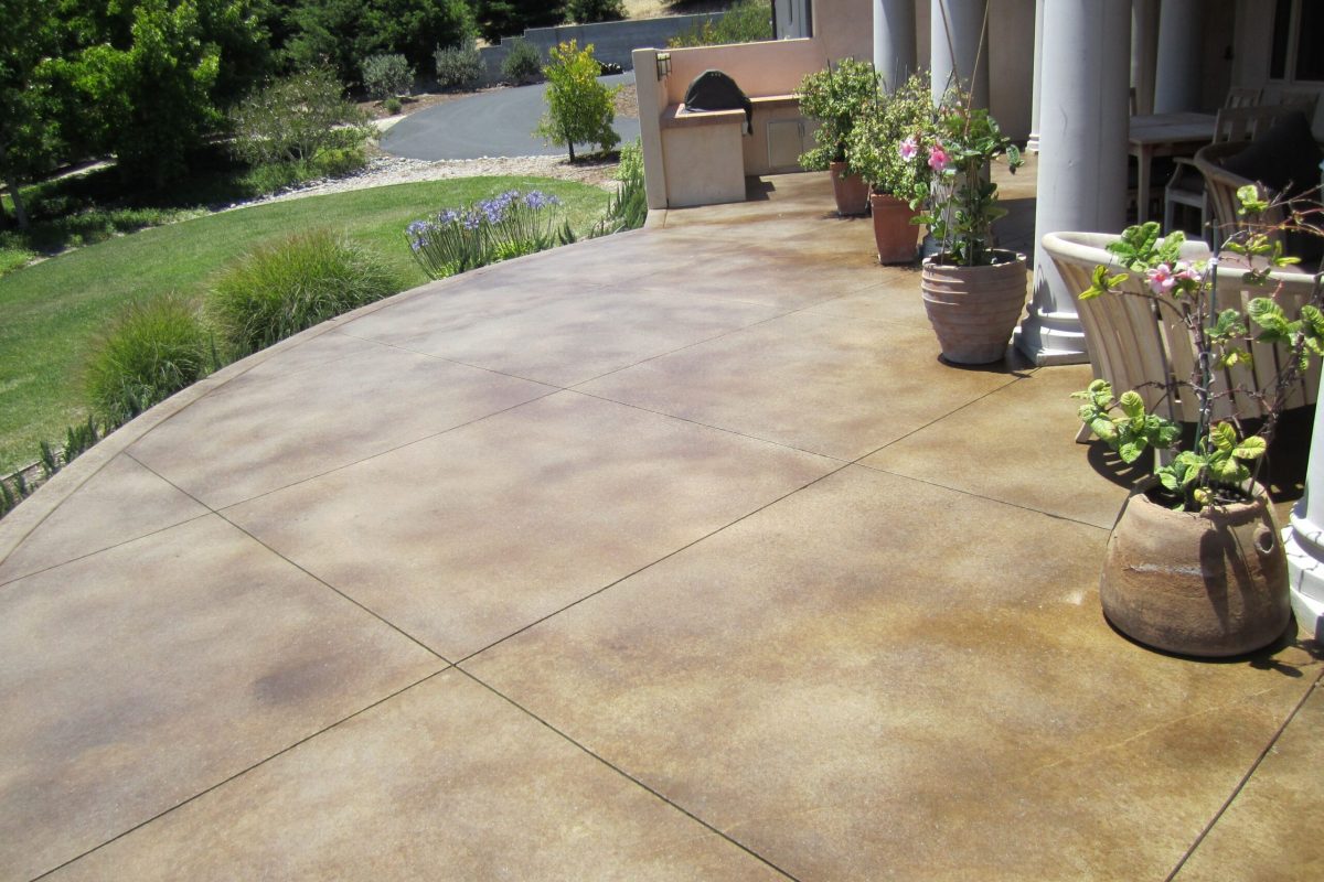 Curved, brown decorative concrete patio with scored lines, potted plants, and a green lawn.