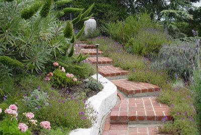 Red brick steps wind up a hillside garden filled with green foliage and pink and purple flowers.