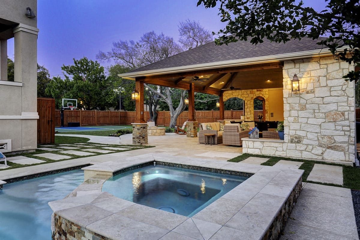 Luxury backyard at dusk with a stone hot tub, a covered lounge area with wicker furniture, and a basketball court.
