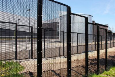 Black metal mesh security fencing enclosing an industrial facility with a large storage tank in the background.