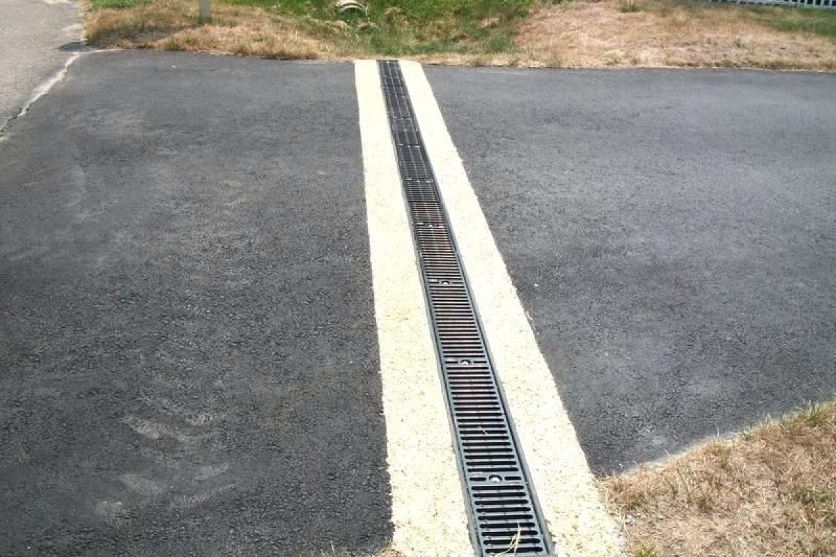 Black metal trench drain grate installed in a concrete strip across a black asphalt driveway.