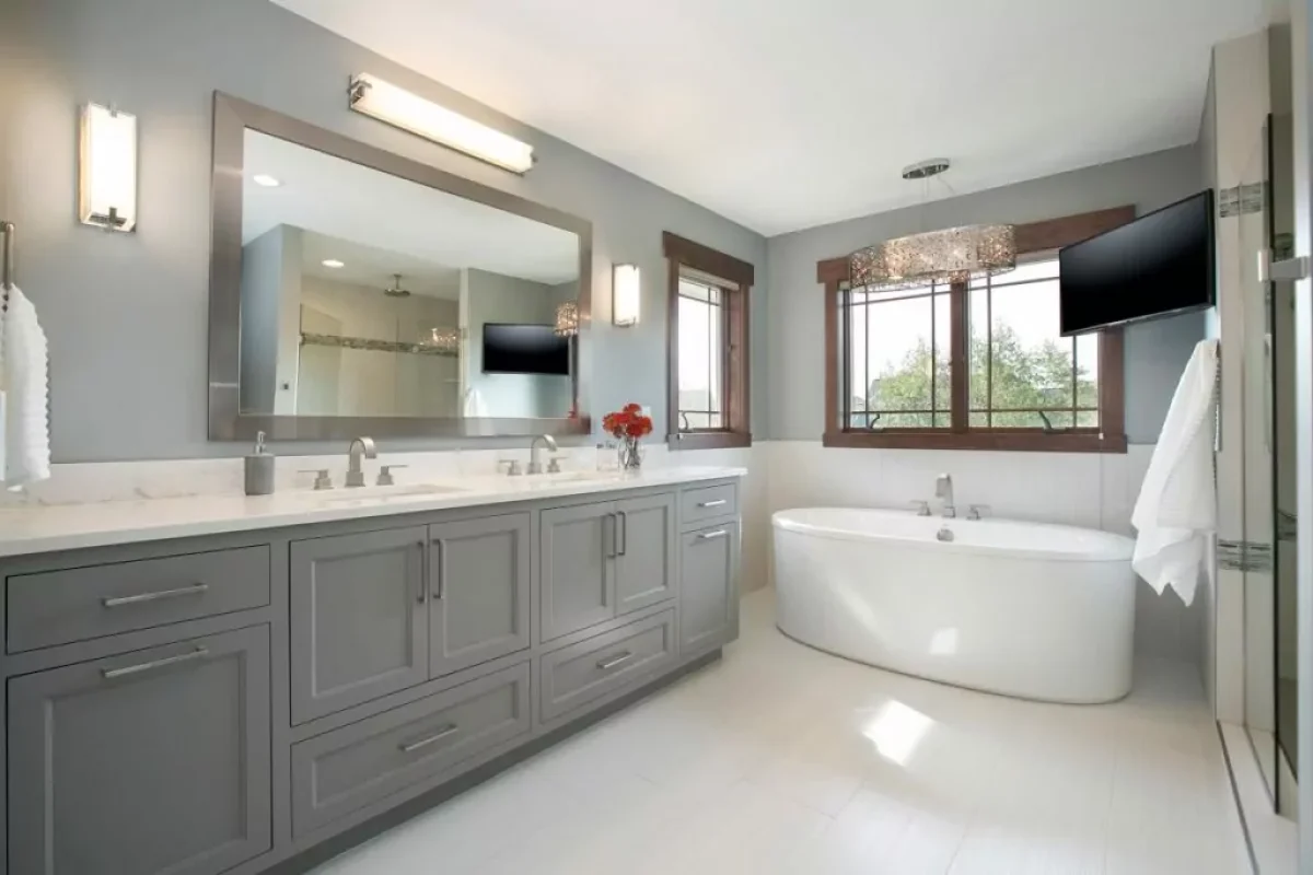 Modern bathroom with grey double vanity, large mirror, freestanding white tub, and a wall-mounted TV.