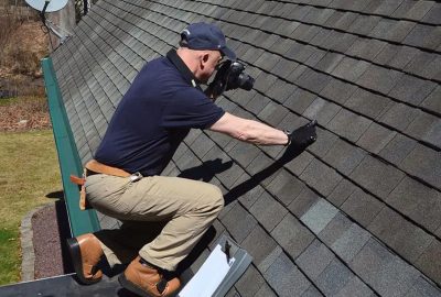 Man kneeling on a shingled roof, taking a photo and pointing to a specific area during an inspection.