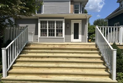 Wide new wooden stairs with white railings leading to a deck at the back of a grey house.