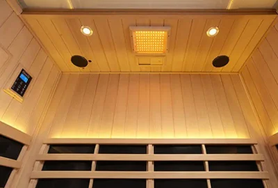 Interior view of a light wood sauna with ceiling lights, speakers, a control panel, and infrared heating panels.