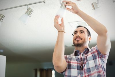 Hispanic man reaches up to install or change light bulb on light fixture in new hoe. He is standing on a ladder and reaching up to the track lighting fixture. He smiles confidently as he works. He is wearing a plaid shirt and has brown hair and a beard.