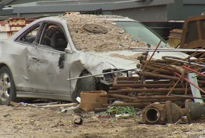 Crushed white car covered in debris and surrounded by scrap metal in a salvage yard.