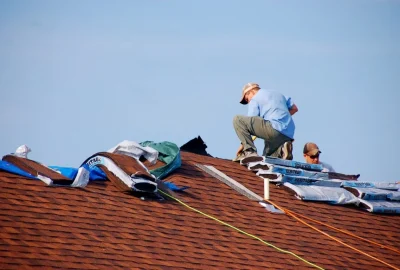 Roofers installing brown shingles on a roof under a clear blue sky.