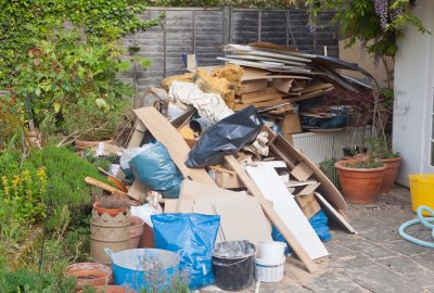 A large pile of construction debris, cardboard, and trash bags sits on a stone patio next to garden plants and pots.