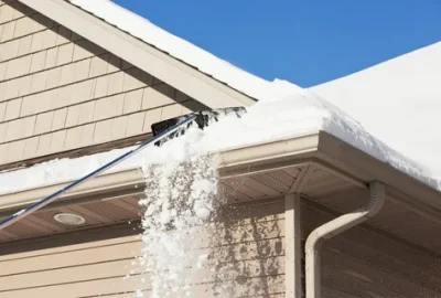 A long-handled tool scrapes snow from a house roof gutter, causing snow to fall.