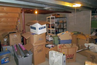 Cluttered storage room with stacks of cardboard boxes, plastic bins, a white fan, and metal shelving.