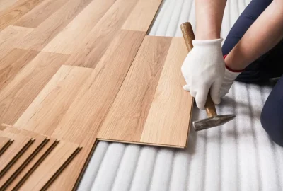 A person wearing gloves using a hammer to install light wood laminate flooring over an underlayment.