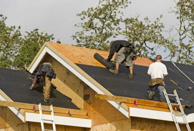 Three construction workers installing black roofing underlayment on a wooden roof structure.