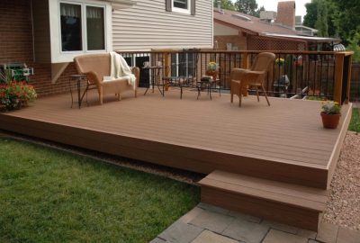 Brown composite deck with wicker furniture, potted plants, and a black railing next to a residential home.