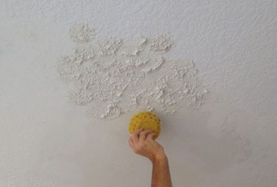 A hand uses a yellow sponge to smooth wet joint compound onto a textured ceiling.