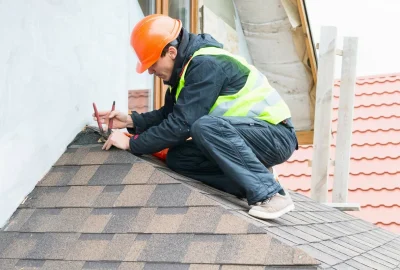 Construction worker in a hard hat and safety vest crouching on a roof to repair asphalt shingles.