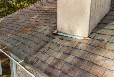 Damaged asphalt shingles and rusted metal flashing around a stucco chimney on a roof with pine needle debris.