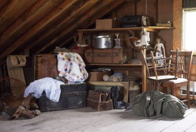 Cluttered wooden attic filled with vintage suitcases, chairs, a patchwork quilt, and antique collectibles.