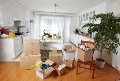 Cardboard boxes of kitchenware scattered across a wooden floor in a white kitchen during a move.