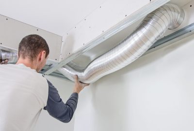 A worker installing flexible metal ventilation ductwork in a ceiling framework.