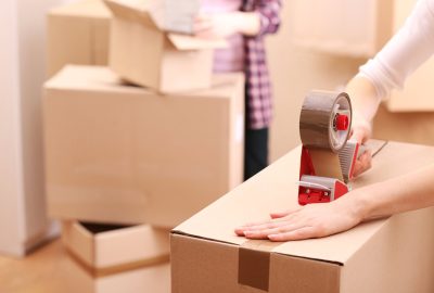 A person uses a red tape dispenser to seal a cardboard moving box.