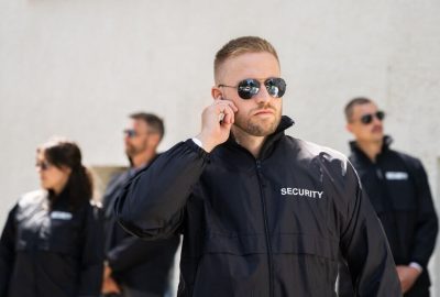 A man in a black security jacket and sunglasses adjusts an earpiece, with other security personnel blurred in the background.
