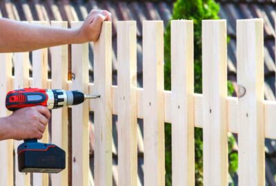 A person uses a cordless power drill to install a wooden picket on a fence.