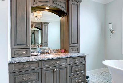 Grey vanity with granite countertop, large mirror, and tall cabinetry next to a white freestanding bathtub.