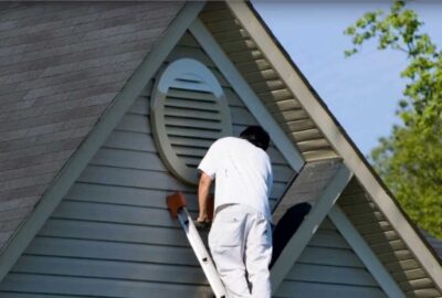 Person on a ladder painting the gable and circular vent of a house.