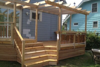 Light-colored wooden deck with wide stairs, railings, and an overhead pergola attached to a blue house.