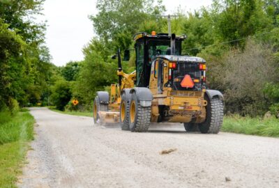 Yellow Caterpillar road grader grading a gravel road in a wooded area.