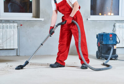 A worker in red overalls uses a heavy-duty industrial vacuum to clean dust from a concrete floor.